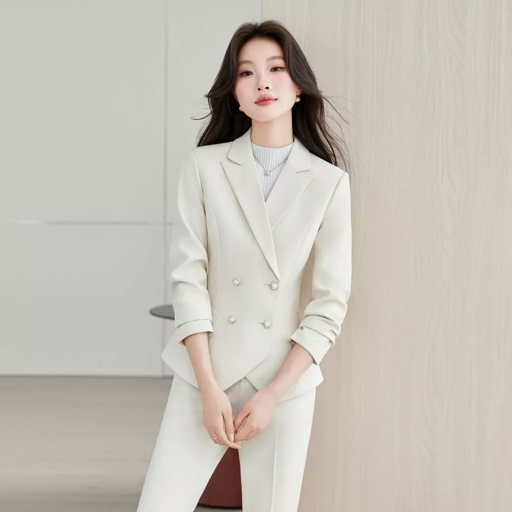 Donna in completo beige con giacca e pantaloni abbinati, appoggiata a parete chiara