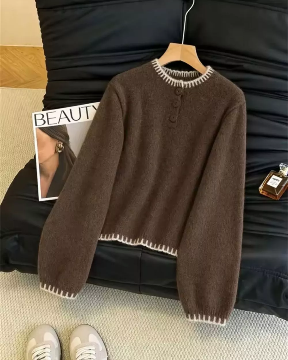 Maglione marrone con dettagli bianchi appoggiato su divano nero con scarpe da ginnastica da donna accanto