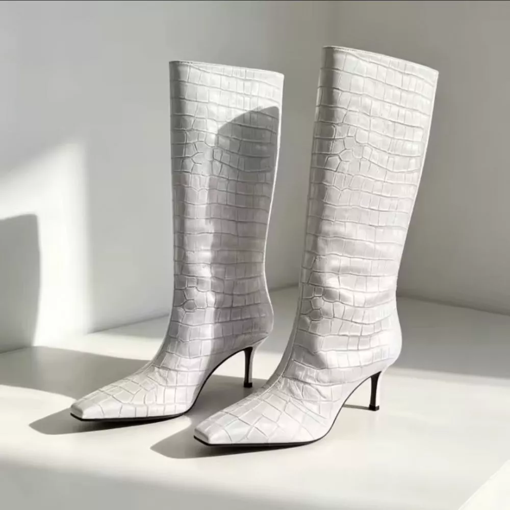 Stivali bianchi sotto al ginocchio in materiale sintetico effetto pelle coccodrillo con tacco sottile visualizzati su sfondo chiaro